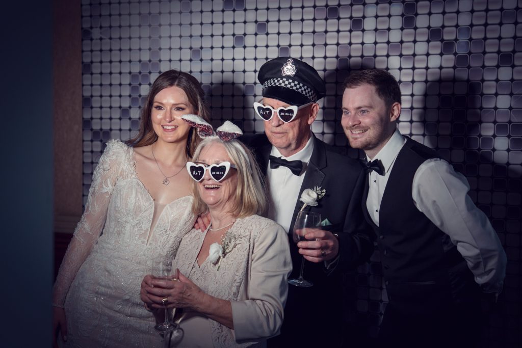 Four people dressed in formal attire at a celebration pose and smile together, wearing novelty heart-shaped sunglasses. The background features a tiled wall with a geometric pattern. Two hold drinks and one wears a police-style hat.