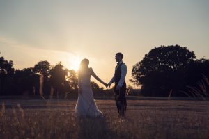 A bride and groom hold hands while facing each other outdoors at sunset, with the sun low in the sky behind them, casting a warm glow and creating their silhouettes against the landscape.