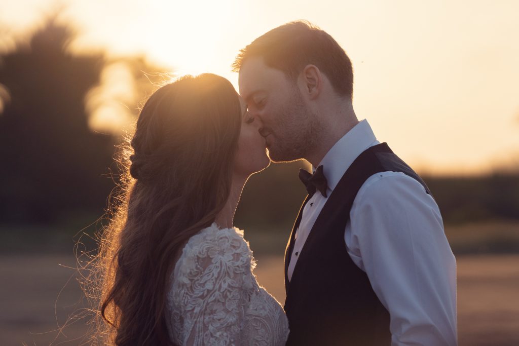 A bride and groom share a romantic kiss at sunset, with warm sunlight glowing behind them. The bride wears a lace dress and has long wavy hair; the groom is in a waistcoat and bow tie.