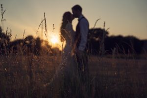 A couple stands close together in a field at sunset, silhouetted against the sun with tall grass in the foreground. The scene feels romantic and serene.