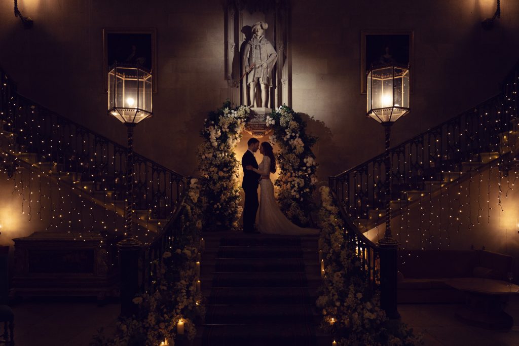 A bride and groom share a kiss on a grand, flower-adorned staircase under soft, romantic lighting, with fairy lights and a large statue in the background.