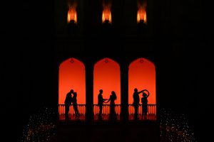 Silhouettes of six people dancing and posing on a balcony with three arches, illuminated by a warm orange-red light, against a dark background with small lights below.