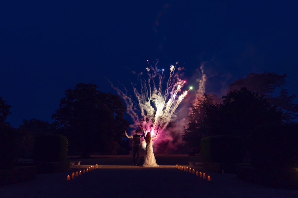 A bride and groom stand hand in hand, watching colourful fireworks light up the night sky, with trees silhouetted in the background and candles lining the pathway towards them.