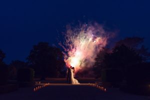 A bride and groom share a kiss at night outdoors, surrounded by lit candles, with colourful fireworks lighting up the dark sky behind them.