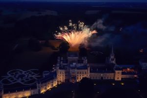 A large, illuminated castle is seen at night with bright fireworks bursting in the sky above the gardens, surrounded by dark trees and open fields in the distance.