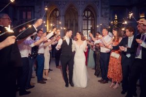 A bride and groom walk hand in hand, smiling, surrounded by guests holding sparklers at night outside a grand stone building with arched windows, celebrating their wedding.