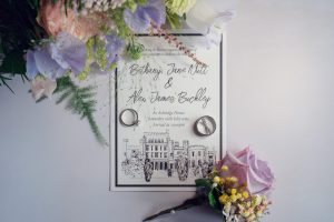 A wedding invitation with two rings placed on it, surrounded by pastel flowers and greenery. The invitation features names, event details, and an illustration of a manor at the bottom.