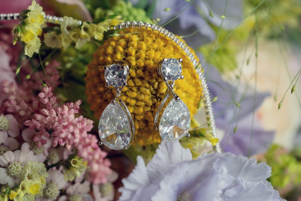 A pair of sparkling diamond earrings rests on a yellow billy ball flower, surrounded by pink, purple, and green flowers and a string of small white pearls.