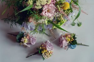 A pastel-coloured floral arrangement with greenery sits above three matching buttonholes on a light surface, featuring roses, yellow billy balls, and assorted delicate flowers.
