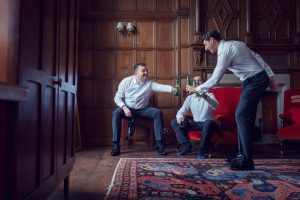 Three men dressed in white shirts and dark trousers are sitting and standing in a wood-panelled room, sharing drinks and toasting bottles while relaxing on red chairs and a patterned rug.