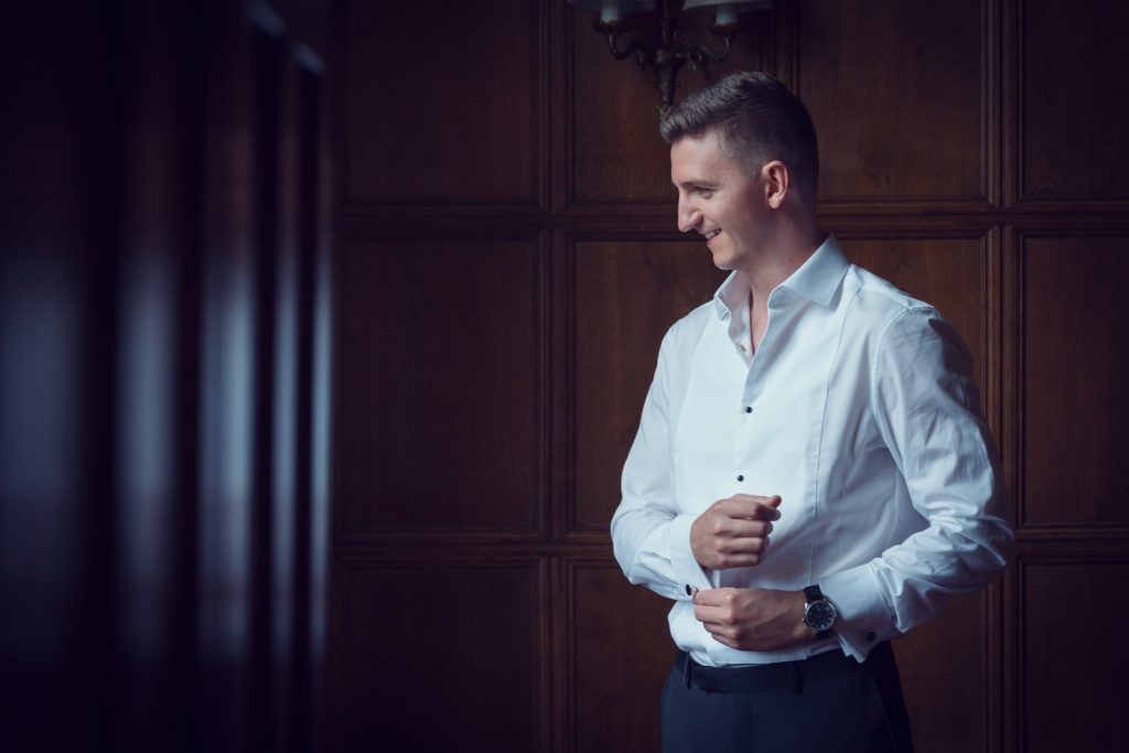 A man in a white dress shirt smiles whilst fastening his cuffs, standing in a dimly lit room with wooden panelled walls.