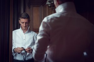 A man stands in front of a mirror, looking down as he prepares to put on a bow tie. He wears a white dress shirt, and another reflection of him is visible from behind. The room has wooden panelling and soft lighting.