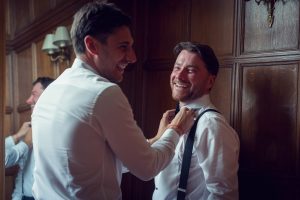 Two men in white shirts laugh as one helps the other adjust his tie in a warmly lit, wood-panelled room. Another smiling person is seen in the background, also adjusting his collar.