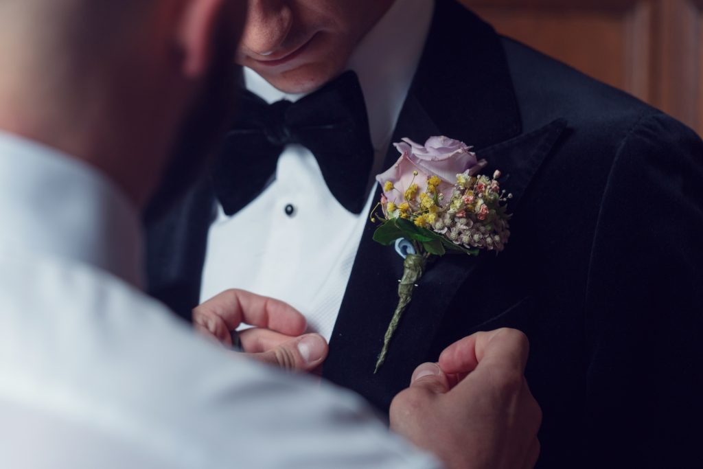 A close-up of someone pinning a colourful buttonhole with pink and yellow flowers onto the lapel of a man wearing a black dinner jacket and bow tie. The focus is on the hands and the buttonhole.