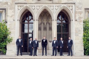 Seven men in dinner jackets stand evenly spaced in front of a large, ornate stone building with tall arched windows and detailed carvings. The group appears formal, posing for a photo outside.