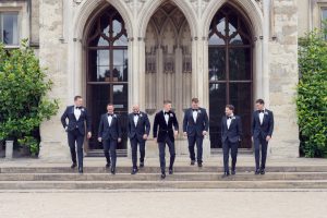 Seven men in formal suits and bow ties walk down stone steps outside a large, historic building with arched windows and detailed stonework, surrounded by greenery.