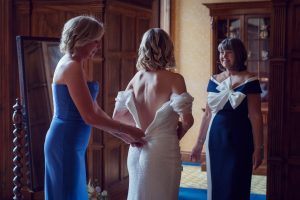 Three women in formal dresses are in a warmly lit room. One woman is fastening the back of another’s white gown, while a third woman watches and smiles, appearing supportive as they prepare for a special event.