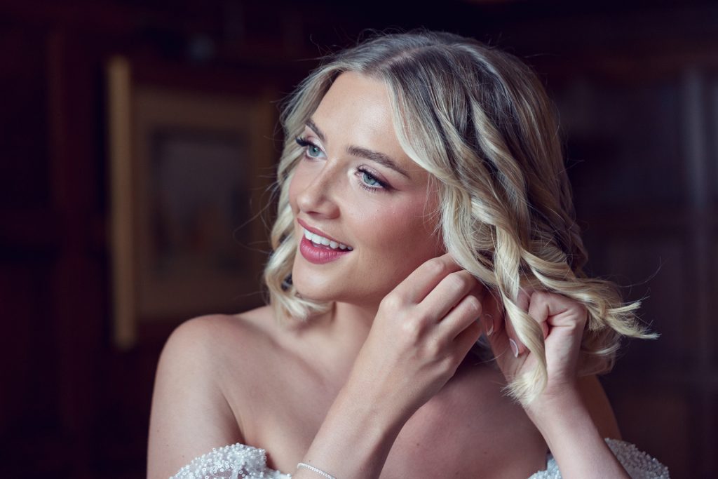 A smiling woman with blonde, wavy hair and light make-up adjusts an earring while wearing an off-the-shoulder white dress in a softly lit, elegant room.