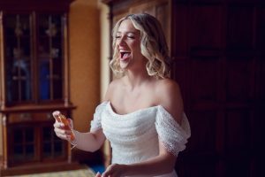 A smiling woman in an off-the-shoulder white dress laughs joyfully whilst holding a small perfume bottle, standing indoors in a warmly lit, elegant room with wooden furniture.