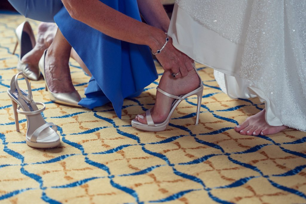 A person in blue helps a bride put on a high-heeled sandal, while another heel sits on the patterned carpet. The bride’s dress and bare foot are visible, suggesting a wedding preparation scene.