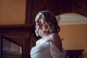 A woman with short, wavy blonde hair wearing an off-the-shoulder, textured white dress stands in a warmly lit, elegant room with wood panelling and an arched ceiling, looking thoughtfully to the side.