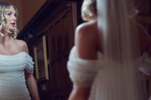 A bride in an off-the-shoulder white dress stands indoors, looking at herself in a mirror. Her blonde hair is styled in loose curls, and a long veil drapes down her back. The setting has dark wooden panelling.