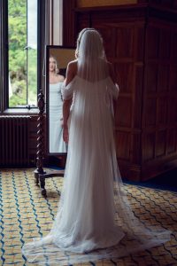 A bride in an off-shoulder white wedding dress and long veil stands facing a mirror in a wood-panelled room, with her reflection showing her face and the front of her dress. Natural light comes in from a window.