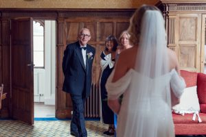 A bride in a white dress stands with her back to the camera as three smiling adults, including an older man in a dinner jacket, look at her with joy in a wood-panelled room.
