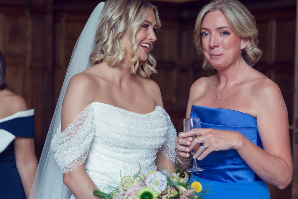 A bride in a white dress smiles whilst standing next to a woman in a blue dress holding a glass, who appears emotional and teary-eyed. Both are indoors, and the bride holds a bouquet.