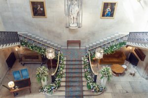 A grand staircase decorated with floral arrangements, featuring patterned carpet and wrought iron railings, in an elegant hall with statues, framed portraits, and plush seating areas on either side.