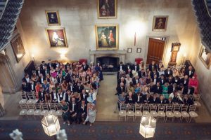 A large group of guests sit in rows of chairs inside a grand, historic room with stone walls, large portraits, and ornate décor, awaiting a ceremony. The room is viewed from above.