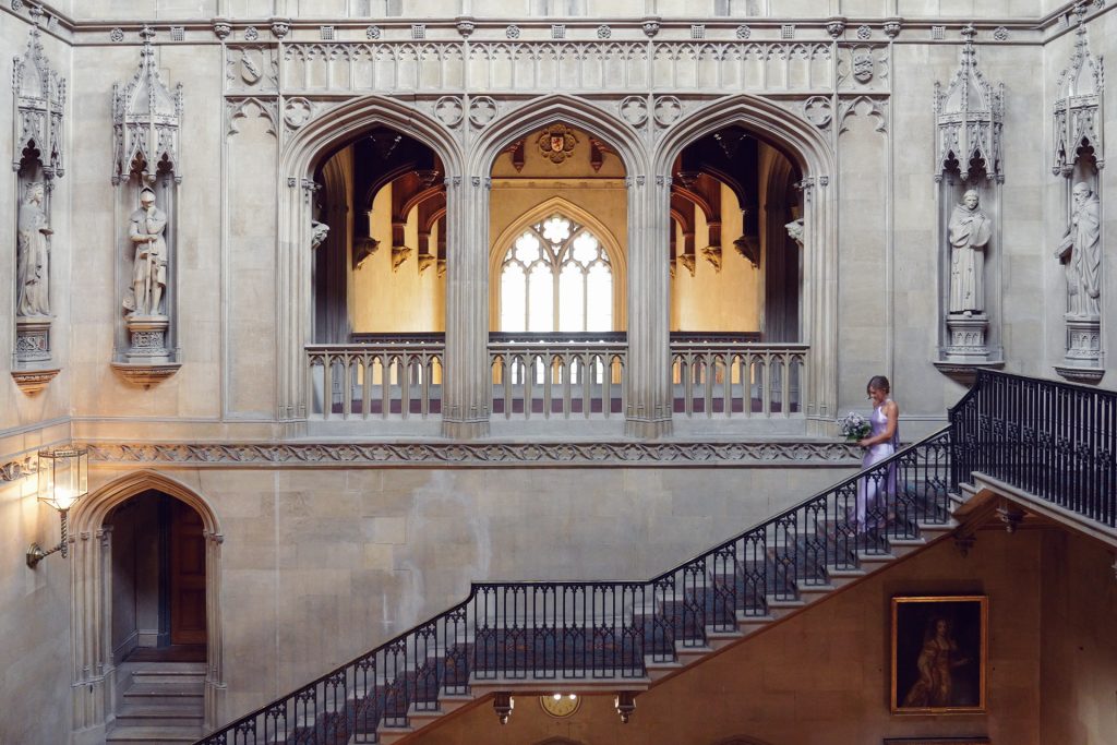 A woman in a lavender dress holding flowers stands on a grand staircase inside a historic stone building with arched windows, statues, and ornate architectural details.