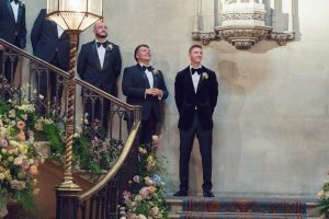 Three men in dinner jackets stand by a staircase decorated with flowers at a formal indoor event, likely a wedding. Two smile and look up, while the third stands behind the bannister. The setting is elegant with ornate architectural details.