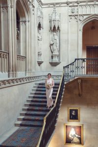 A bride in a white dress and glasses holds a bouquet while descending an ornate staircase in a grand, historic building with statues, arched doorways, and framed portraits on the stone walls.