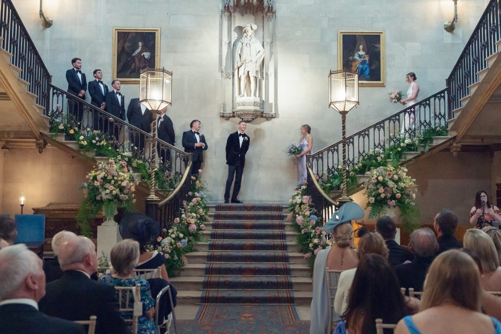 A bride and groom stand on a grand staircase, surrounded by their wedding party. Guests are seated below. The venue is elegant with floral arrangements, large paintings, and a statue at the top of the stairs.