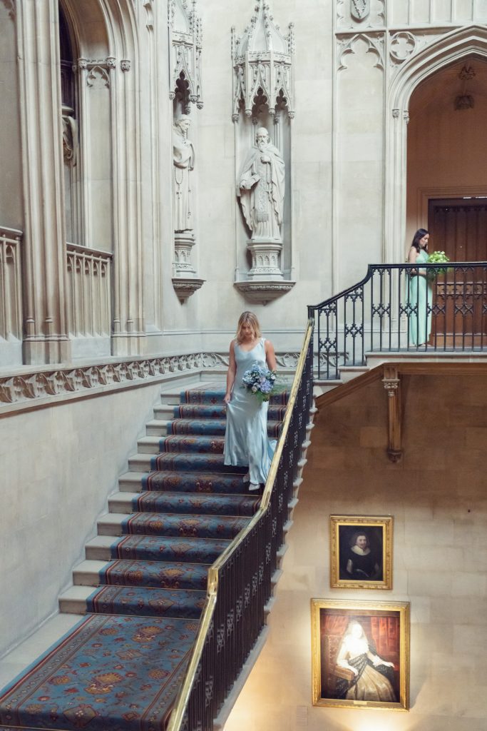Two women in light dresses walk down an ornate stone staircase with a patterned carpet, inside a grand building adorned with statues and framed portraits on the walls. One woman holds a bouquet of flowers.
