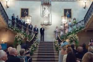 A bride and groom stand at the top and centre of a grand staircase, flanked by their wedding party. Guests are seated below, and the staircase is decorated with lush floral arrangements. The setting is elegant and formal.