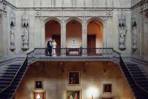 A bride and groom stand together on an ornate balcony inside a grand, historic building with arched stone details, statues, and portrait paintings on the walls beneath them.