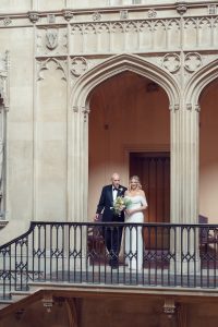 A bride in a white dress and veil stands beside an older man in a suit, holding a bouquet, on a grand balcony with ornate stone arches and carvings in an historic building.