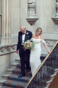 A bride in an off-the-shoulder white gown holds a bouquet and smiles as she walks down a grand staircase with an older man in a tuxedo, inside an ornate, historic building.