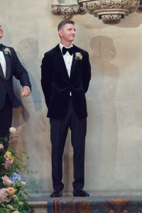 A man in a black dinner jacket with a bow tie and buttonhole stands indoors on a stone floor, hands behind his back, near a floral arrangement, looking slightly to his left and smiling.