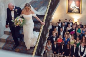 A bride in a white dress walks down a grand staircase with an older man, likely her father, while guests in formal attire watch from below in an elegant venue.