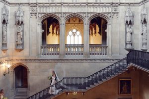 A bride in a white dress and veil walks up a grand stone staircase in an historic building with ornate arches, statues, and large windows. Another person stands at the bottom of the stairs.