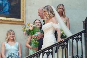 A smiling bride in an off-the-shoulder white gown and veil walks down the stairs, holding a bouquet, accompanied by an older man. Bridesmaids in pastel dresses stand behind them, holding flowers.
