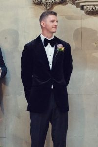 A man in a black dinner jacket with a bow tie and a flower buttonhole stands against a stone wall, looking to the side with his hands behind his back.