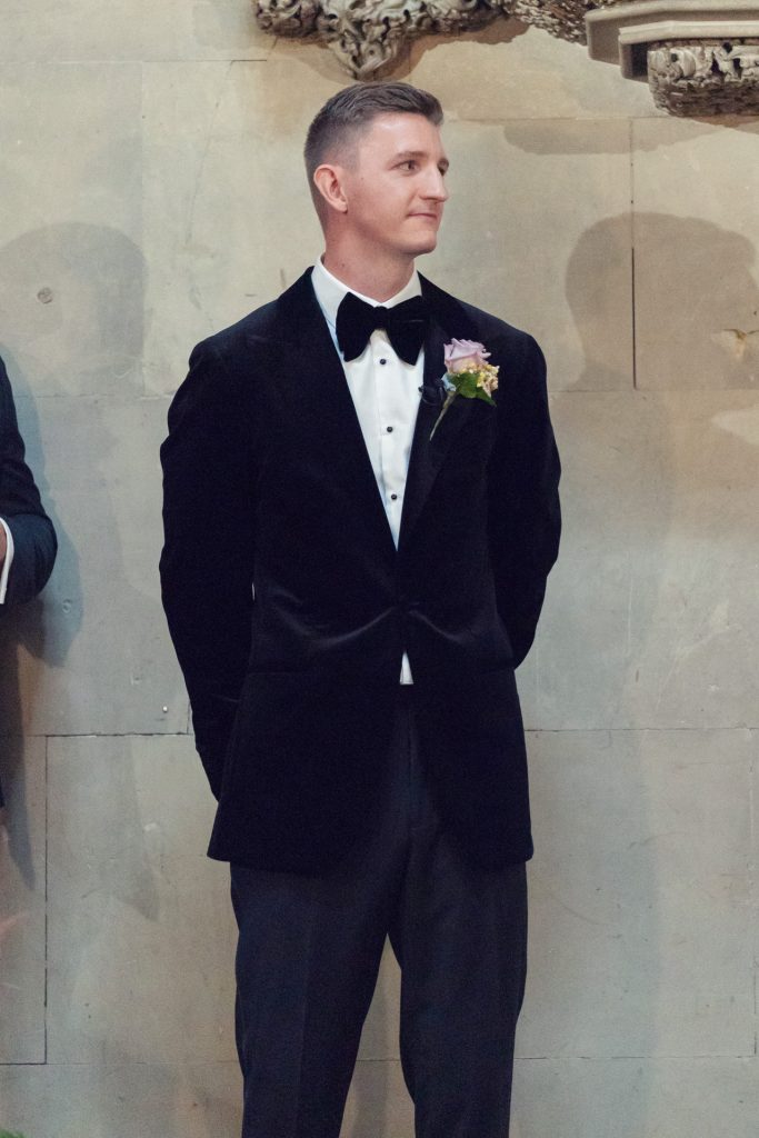 A man in a black dinner jacket with a bow tie and a flower buttonhole stands against a stone wall, looking to the side with his hands behind his back.