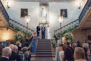 A wedding ceremony inside a grand hall with a couple and an officiant standing on stairs, surrounded by flowers and guests seated, ornate railings, large paintings, and a statue in the background.