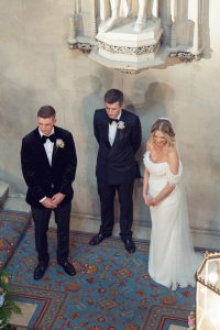 Three people stand indoors on a patterned carpet during a formal ceremony. Two men wear black dinner jackets with buttonholes, and a woman in a white off-the-shoulder wedding dress stands beside them, all facing forward with heads slightly bowed.