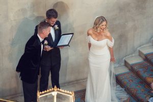 A bride in a white dress laughs joyfully on carpeted stairs beside two men in suits, one holding a folder, against a stone wall, during a wedding ceremony.