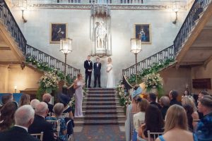 A wedding ceremony in an elegant hall with a central staircase; a couple stands with an officiant at the top of the stairs, surrounded by flowers, whilst guests watch from below.